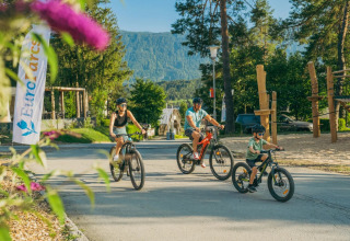Familie cykler ved et feriecenter med glamping, legeplads og skovklædte bjerge i baggrunden.