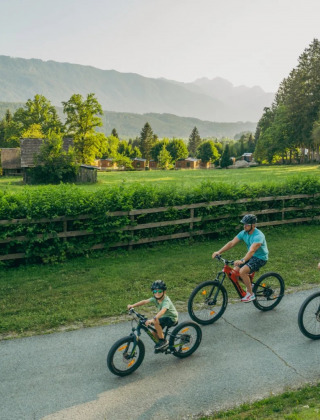 Gezin fietst over pad bij vakantiepark met glamping, groene velden en bergen op de achtergrond.