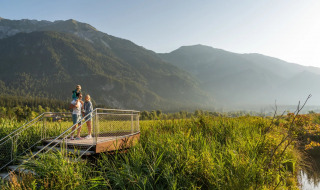 Gezin op uitkijkplatform in een vakantiepark, omgeven door groene velden en bergen op de achtergrond.