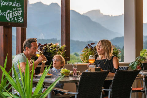 Family enjoying dinner and drinks on a glamping park terrace with scenic mountain views behind.