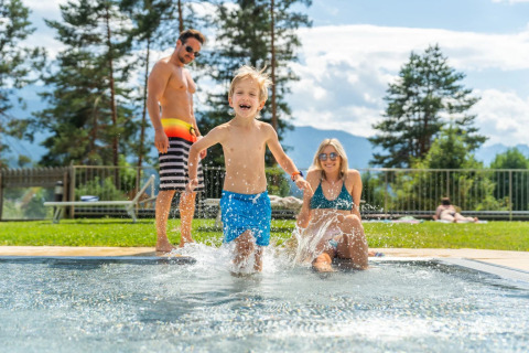 Happy family enjoys a summer day at a holiday park pool with glamping, surrounded by trees and mountains.