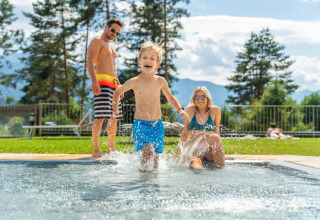 Happy family enjoys a summer day at a holiday park pool with glamping, surrounded by trees and mountains.