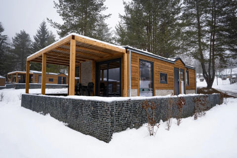 Bungalow moderno in legno con veranda coperta in un parco vacanze innevato, circondato da pini.