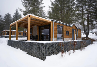 Cabaña moderna de madera con terraza cubierta en parque vacacional nevado, rodeada de árboles altos.