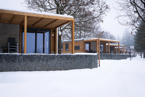 Modern glamping cabins with wooden terraces and stone fences surrounded by fresh snow at a holiday park.