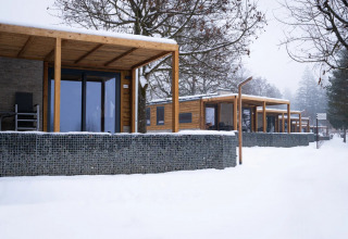 Glampinghytter med træterrasser og stenhegn dækket af sne i et ferieparklandskab om vinteren.