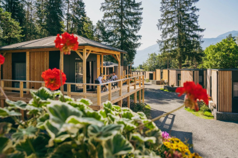 Parco vacanze con alloggi glamping in legno, tra fiori e pini, persone che si rilassano sulle verande.