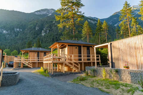 Bungalow in legno per glamping in un villaggio vacanze tra montagne verdi e pini, al tramonto.