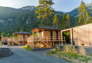 Holzhütten auf einem Glamping-Ferienpark, umgeben von Bergen und Wald, im sanften Abendlicht.