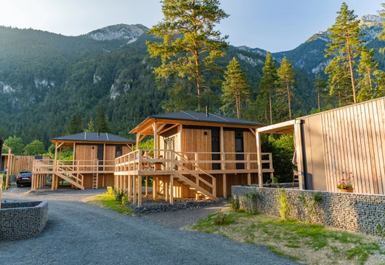 Holzhütten auf einem Glamping-Ferienpark, umgeben von Bergen und Wald, im sanften Abendlicht.