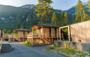 Wooden glamping cabins at a holiday park, nestled in majestic mountains and tall pine trees at sunset.