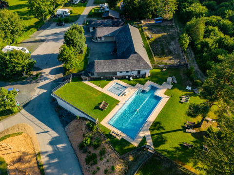 Vista aérea de un parque vacacional con piscina grande, césped verde y zonas de glamping rodeadas de naturaleza.