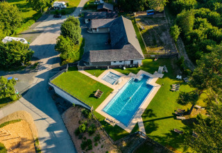Vista aérea de un parque vacacional con piscina grande, césped verde y zonas de glamping rodeadas de naturaleza.