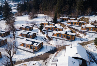 Luftfoto af sneklædt feriepark med glampinghytter, omgivet af træer og bjerge om vinteren.