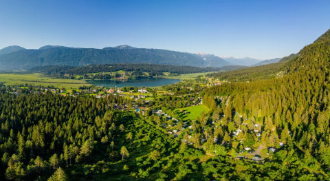 Vista aerea di un villaggio vacanze con sistemazioni glamping tra bosco, lago e montagne sullo sfondo.
