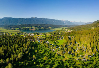Luftfoto af feriepark med glamping telte blandt skov, sø og bjerge under blå himmel.