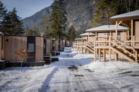 Fila di moderne cabine glamping in un parco vacanze innevato, circondato da montagne e pini in inverno.