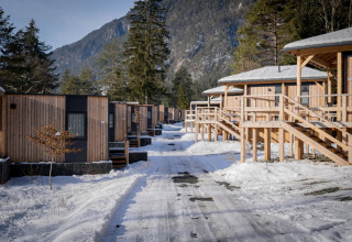 Fila de cabañas glamping modernas en un parque de vacaciones nevado, rodeado de montañas y pinos en invierno.