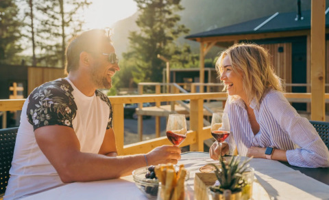 Pareja disfrutando vino al aire libre en una mesa bajo el sol, rodeados de cabañas en un parque glamping.