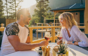 Par nyder vin udendørs ved et bord i solen, omgivet af træhytter i et glamping-område.
