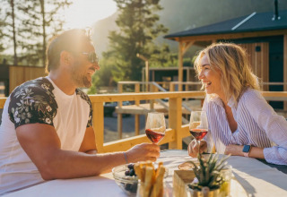 Par nyder vin udendørs ved et bord i solen, omgivet af træhytter i et glamping-område.