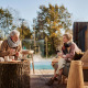Ældre par nyder vin og snacks udendørs ved Unbrick One lodge med sauna og pool i baggrunden.
