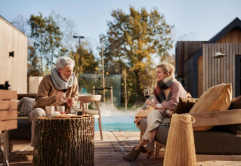 Senior couple enjoying wine and snacks outdoors at Unbrick One lodge with sauna and pool in view.