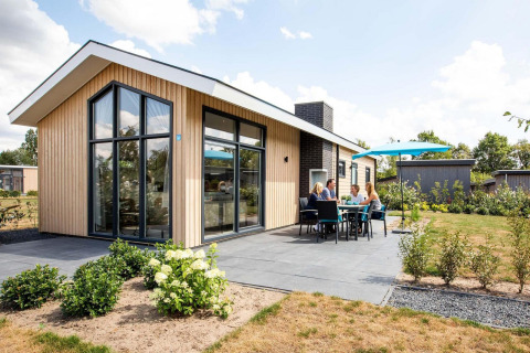 Moderno lodge Berkenhoeve con grandi finestre, patio e persone che pranzano all'aperto sotto un ombrellone blu.