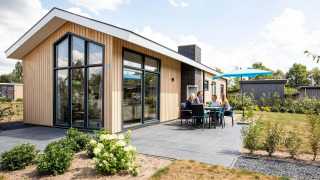 Moderna cabaña Berkenhoeve con grandes ventanales, terraza y personas disfrutando de comer al aire libre bajo una sombrilla azul.