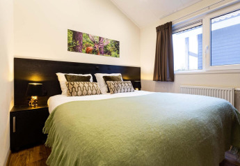 Dormitorio en Berkenhoeve Wellness lodge con cama doble, manta verde y ventanas grandes iluminadas.
