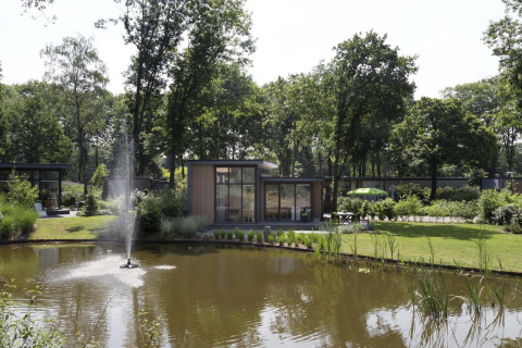 Una elegante cabaña junto a un estanque con fuente en Landgoed de Scheleberg, Países Bajos, rodeada de árboles.