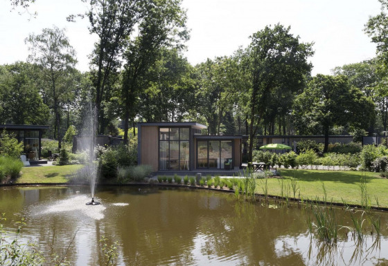 Una elegante cabaña junto a un estanque con fuente en Landgoed de Scheleberg, Países Bajos, rodeada de árboles.