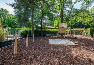 Playground with a sandpit, wooden playhouse, swings and slide surrounded by trees at Het Verscholen Dorp park.