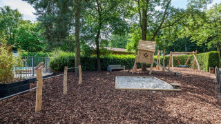 Zona de juegos con arenero, columpios, tobogán y cabaña rodeada de árboles en Het Verscholen Dorp.