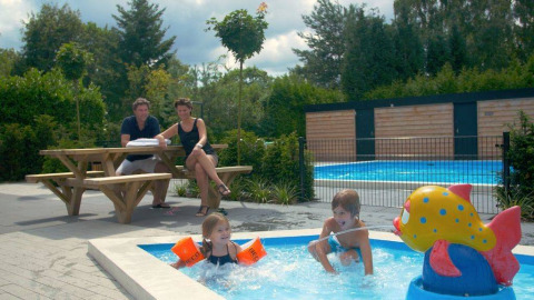 Familia sentada en mesa de picnic, niños juegan en piscina infantil con fuente en Het Verscholen Dorp.