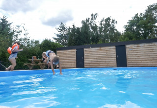 To børn leger ved en swimmingpool i en feriepark med træhegn og grønne træer i Bungalowpark Het Verscholen Dorp.