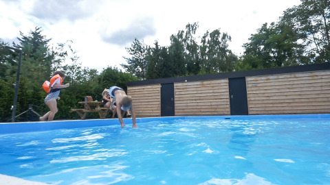 To børn leger ved en swimmingpool i en feriepark med træhegn og grønne træer i Bungalowpark Het Verscholen Dorp.