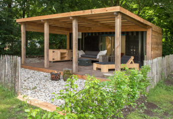 Udendørs opholdsområde ved tiny house ‘Sea container’ med træterrasse, sofaer og grønt hegn.