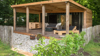 Udendørs opholdsområde ved tiny house ‘Sea container’ med træterrasse, sofaer og grønt hegn.