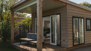 Cozy wooden lodge with a covered porch, outdoor seating, and large windows, surrounded by greenery.