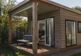 Accogliente lodge in legno con veranda coperta, divanetto all'aperto e grandi finestre in mezzo al verde.
