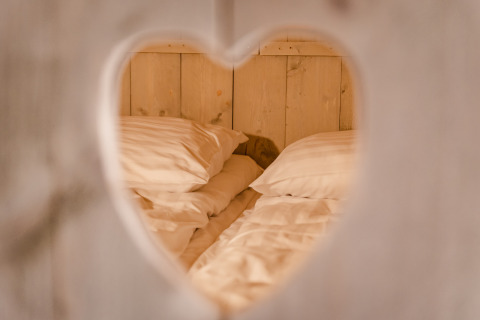 View through a heart-shaped cutout reveals a cozy bed with white pillows at Glamping Lodge, Netherlands.