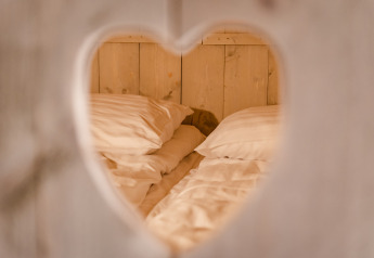 View through a heart-shaped cutout reveals a cozy bed with white pillows at Glamping Lodge, Netherlands.