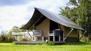 Imagen de una Tiny Tent lodge con porche y sillas sobre césped verde bajo un cielo parcialmente nublado.