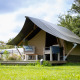 Imagen de una Tiny Tent lodge con porche y sillas sobre césped verde bajo un cielo parcialmente nublado.