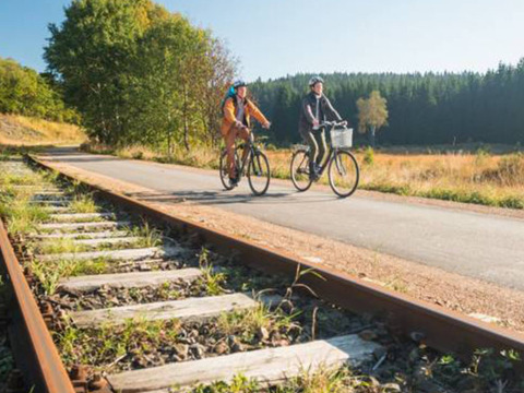 Two cyclists ride on a paved trail beside old railway tracks at Camping Oos Heem in Liège, Belgium.