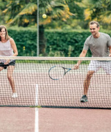 Zwei Erwachsene spielen lachend Tennis auf einem Außenplatz mit Palmen und grünen Hecken im Hintergrund.