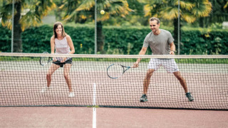 Zwei Erwachsene spielen lachend Tennis auf einem Außenplatz mit Palmen und grünen Hecken im Hintergrund.