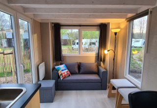 Bright living room inside a tiny house at Vakantiepark 't Urkerbos, Netherlands, with sofa, windows and outdoor view.