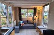 Salon lumineux d'une tiny house au Vakantiepark 't Urkerbos, Pays-Bas, avec canapé, grandes fenêtres et vue extérieure.